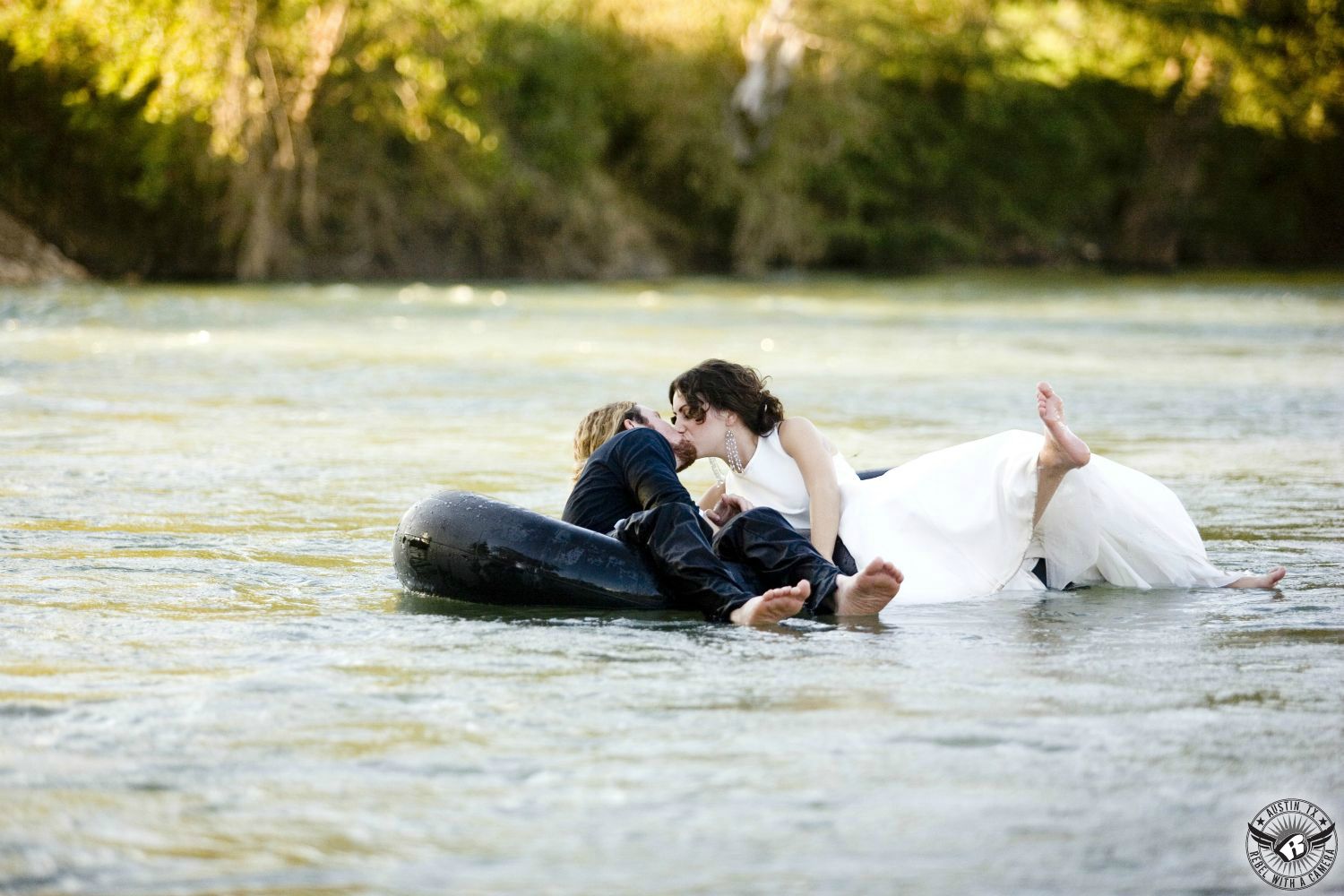 Dark haired girl wears mother wedding dress barefoot floating down the river in a tube kissing a blond guy wearing a tux shirt and black pants also barefoot and also in an inner tube on the Guadalupe River with green trees in the background in this lighthearted summer engagement picture near Bastrop  